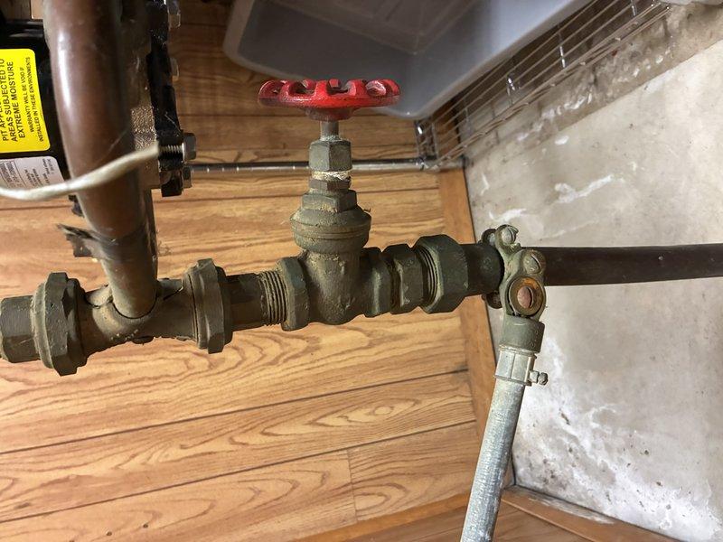 Inspected aging plumbing system with focus on manual gate valve installation. Examined copper and brass pipe assembly with red control valve handle showing signs of wear. Verified valve functionality and checked connections for leaks at threaded joints where components meet the wood-paneled wall.