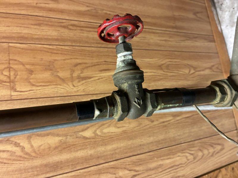 Inspected aging plumbing system with focus on manual gate valve installation. Examined copper and brass pipe assembly with red control valve handle showing signs of wear. Verified valve functionality and checked connections for leaks at threaded joints where components meet the wood-paneled wall.