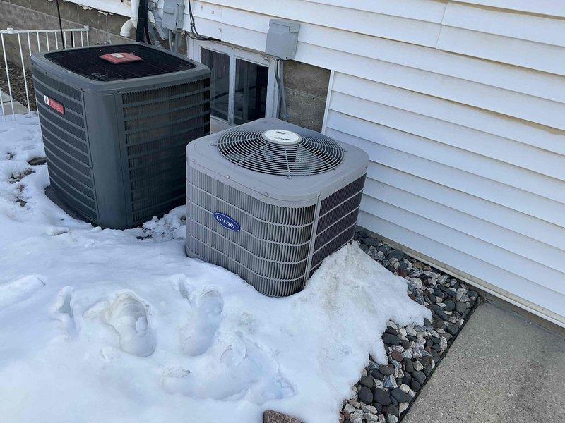 Completed installation of a Carrier air conditioning system with dual outdoor condensers positioned on a snow-covered ground adjacent to the client's vinyl-sided residence. The installation included proper electrical connections and refrigerant line setup. In the basement utility room, installed a new furnace unit with complete PVC ventilation system and organized refrigerant line connections. All components were properly secured and tested for functionality.