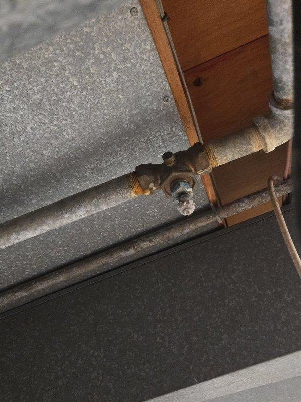 Inspected plumbing system and identified significant corrosion and oxidation on metal pipe fittings, particularly at T-junction/valve assembly and connection points. Documented extensive rust formation on aging pipes in utility area, with deterioration most severe at junction points.