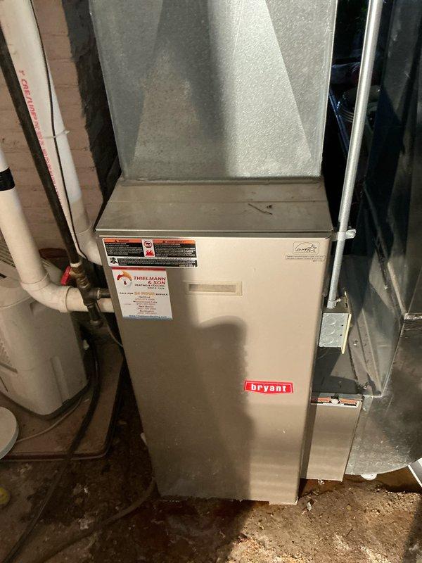 Inspected Bryant furnace unit installed in basement utility area. Unit appears functional with proper ductwork connections and PVC venting. Surrounding area contains numerous stored items that could potentially restrict airflow and access. Recommended replacement due to age and efficiency concerns, with proper clearance of storage items around new installation.