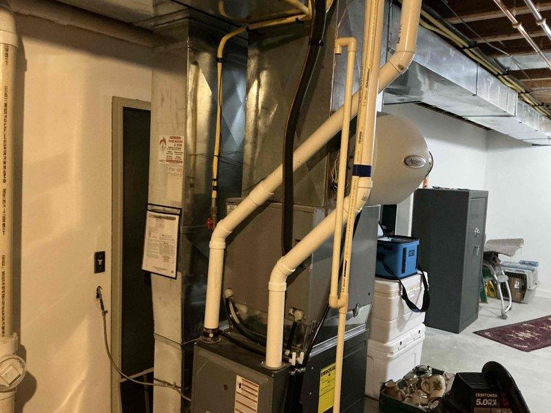 Performed comprehensive inspection and documentation of existing HVAC system in residential basement. System includes furnace unit with sheet metal housing connected to vertical PVC ventilation pipes, side-mounted humidifier, and rectangular metal ductwork running along ceiling joists. Photos captured both the main mechanical room setup and adjacent storage area with exposed infrastructure for complete system evaluation prior to replacement estimate.