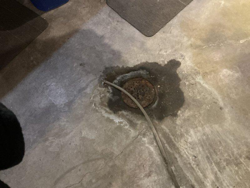 Completed inspection of floor drain system showing significant corrosion and clogging issues. Identified rusted drain cover with debris accumulation and surrounding concrete deterioration. Assessed exterior PVC vent pipe mounted on brick wall for proper installation and functionality as part of the drainage system replacement evaluation.