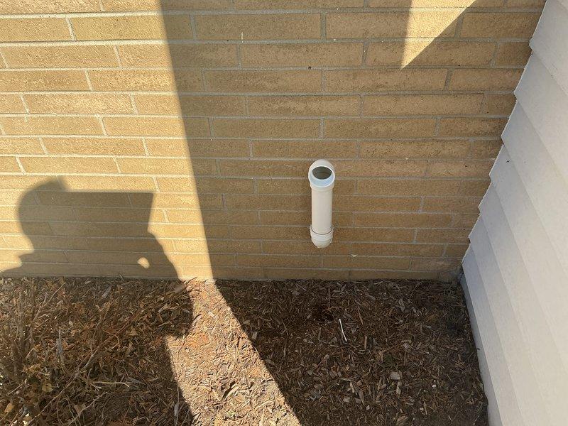 Completed inspection of floor drain system showing significant corrosion and clogging issues. Identified rusted drain cover with debris accumulation and surrounding concrete deterioration. Assessed exterior PVC vent pipe mounted on brick wall for proper installation and functionality as part of the drainage system replacement evaluation.