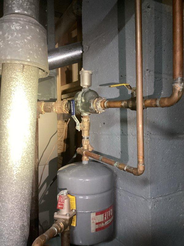 Inspected and documented existing boiler system including expansion tank with proper labeling and associated copper piping network. System features multiple valve assemblies and pressure regulation components mounted to concrete block wall in utility area. All connections appear intact with expected aging patina on copper components.
