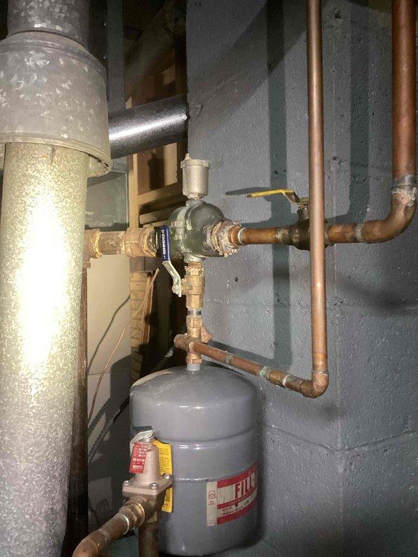 Inspected and documented existing boiler system including expansion tank with proper labeling and associated copper piping network. System features multiple valve assemblies and pressure regulation components mounted to concrete block wall in utility area. All connections appear intact with expected aging patina on copper components.