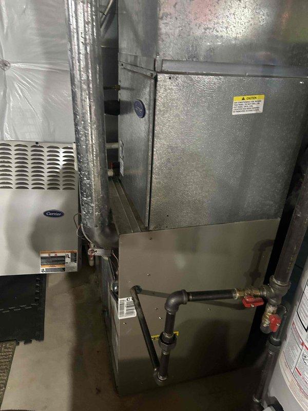 Completed installation of a complete home HVAC system in the utility room, including a Carrier air conditioning unit and metal furnace with proper ductwork connections. System features galvanized steel air handler/plenum, PVC drainage, copper refrigerant lines, and gas connections, all professionally installed alongside the existing water heater.