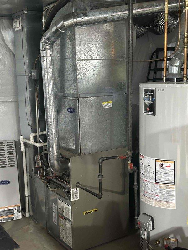 Completed installation of a complete home HVAC system in the utility room, including a Carrier air conditioning unit and metal furnace with proper ductwork connections. System features galvanized steel air handler/plenum, PVC drainage, copper refrigerant lines, and gas connections, all professionally installed alongside the existing water heater.