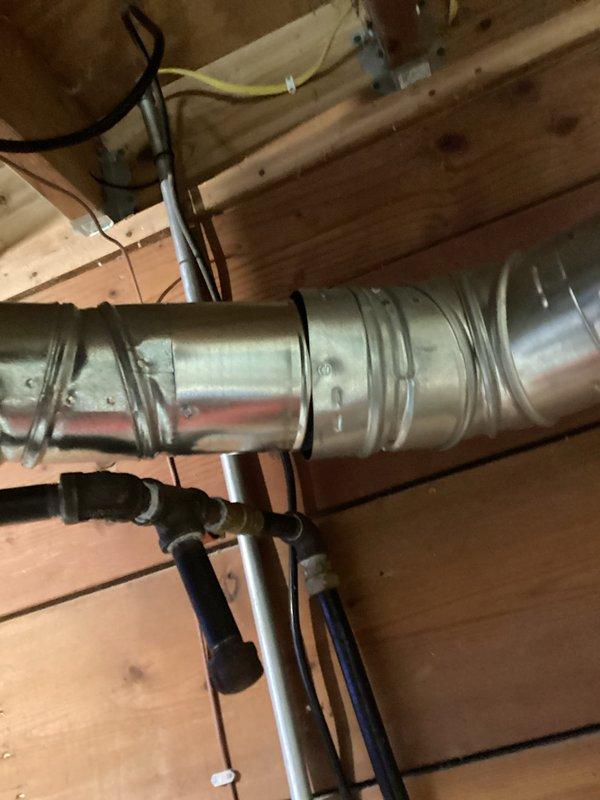 Inspected and serviced wall-mounted HVAC unit in workshop area, verifying proper connection of ductwork to system. Confirmed all seams and joints in metal ductwork were secure and correctly installed through exposed joists, with particular attention to the curved section and connection points to smaller conduits.