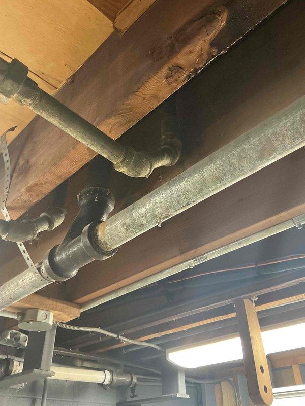 Conducted comprehensive inspection of exposed plumbing system in unfinished ceiling area, specifically examining the cast iron water service pipes showing signs of aging and discoloration. Documented multiple horizontal pipe runs with various connection points and fittings mounted along wooden joists, noting areas of potential concern including corrosion and aging joints that may require future replacement to prevent water damage.