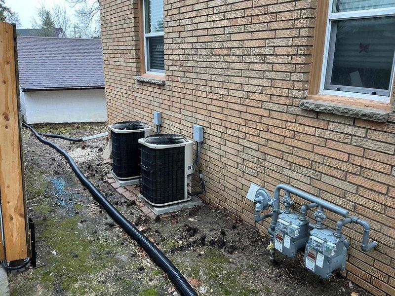 Conducted site inspection of dual outdoor condensing units requiring replacement, documenting their current installation on concrete pads adjacent to gas meters and drainage infrastructure. Interior assessment of mechanical room revealed integration with existing water heater system and associated plumbing, with particular attention to connection points requiring modification during HVAC system replacement.