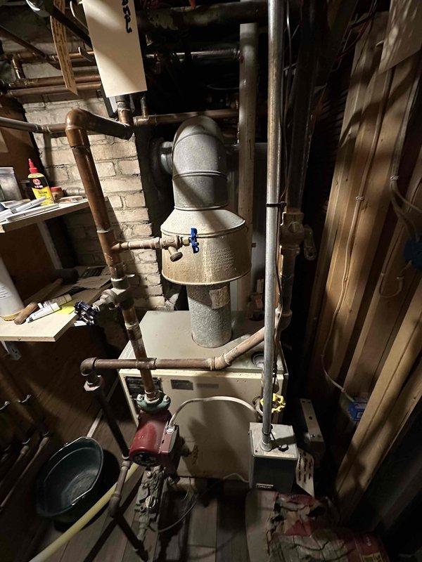 Performed inspection of existing boiler system and electrical panel. Documented current installation showing aging cylindrical water heater/boiler with conical top section connected to complex copper piping network in basement utility area. Electrical service panel appears in good working condition with proper labeling. Noted sump pump system with red motor component installed nearby. Photographs taken of all components to support comprehensive estimate for recommended system upgrades.
