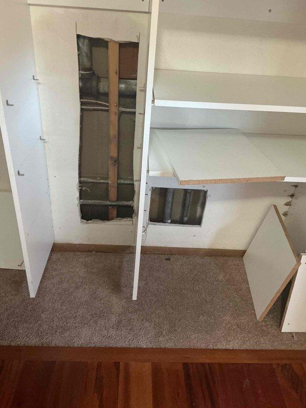 Completed wall access through built-in cabinetry to expose plumbing infrastructure. Created rectangular cutout revealing wall cavity with studs, pipes, and ductwork for necessary repairs. Maintained careful approach to preserve surrounding cabinetry structure during service work.