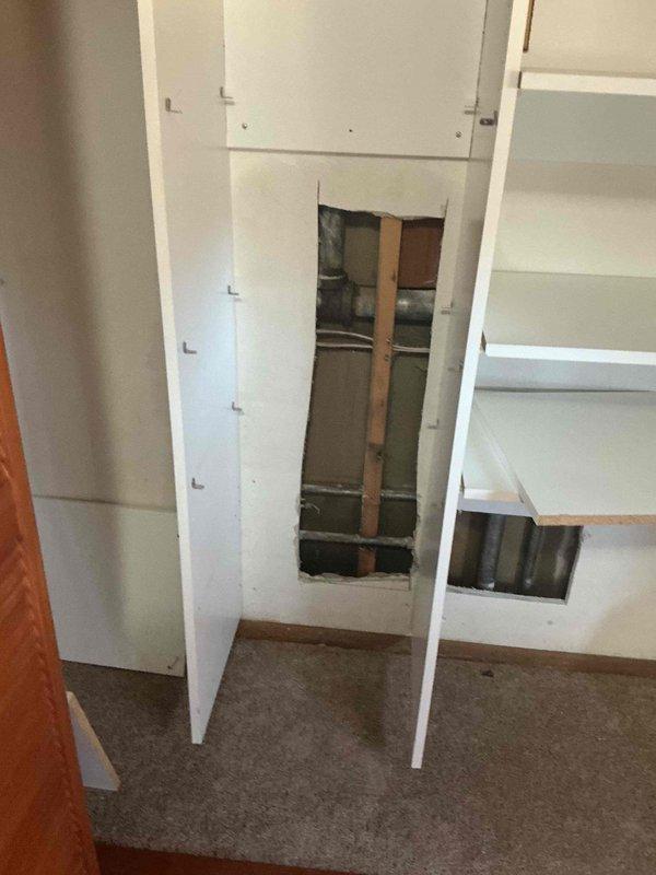 Completed wall access through built-in cabinetry to expose plumbing infrastructure. Created rectangular cutout revealing wall cavity with studs, pipes, and ductwork for necessary repairs. Maintained careful approach to preserve surrounding cabinetry structure during service work.