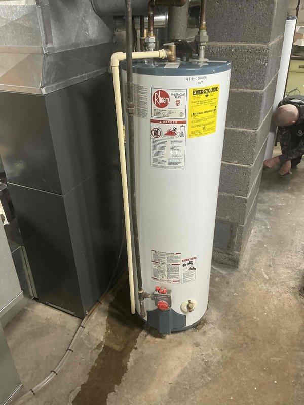 Inspected existing Rheem water heater installation in utility room showing signs of minor leakage at base with visible water staining on concrete floor near drain. Unit appears functional but aging, with standard connections to copper and PVC piping. Recommended replacement of water heater to prevent potential future water damage and ensure continued reliable hot water service.