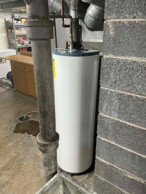 Inspected existing Rheem water heater installation in utility room showing signs of minor leakage at base with visible water staining on concrete floor near drain. Unit appears functional but aging, with standard connections to copper and PVC piping. Recommended replacement of water heater to prevent potential future water damage and ensure continued reliable hot water service.