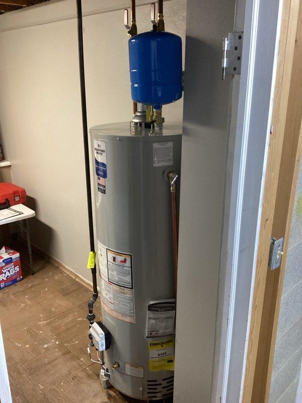 Completed installation of a new residential water heater system with proper connections to existing plumbing. Installed blue expansion tank with copper pipes and brass valves on top of the unit, and ensured pressure relief valve was correctly positioned. System was tested for proper operation and all connections were verified leak-free before completing the job.