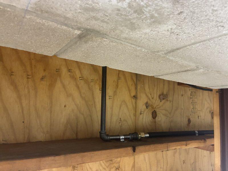 Installed new gas piping system throughout the interior structural space, including horizontal black pipe with elbow joints connecting to vertical sections. Additionally, installed an exterior water valve fixture with red handle and blue center marking on the building's exterior white siding, ensuring proper connection to the main supply line.