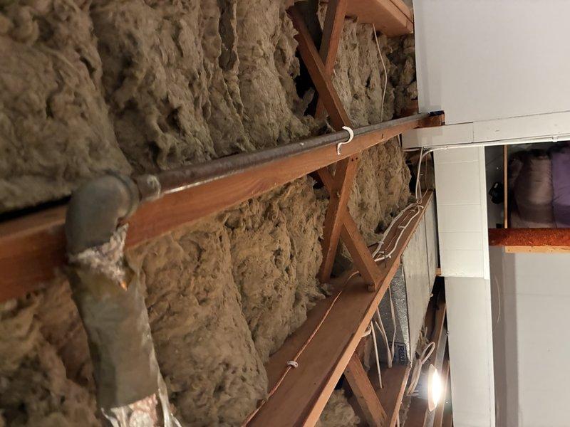 Conducted inspection of residential property to provide piping estimate, documenting exterior access point via wooden deck stairs and examining interior wall cavity. Identified existing plumbing infrastructure with horizontal pipe runs through wooden framing members and assessed insulation condition surrounding piping system for potential replacement or modification work.
