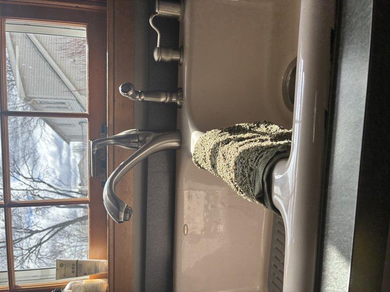 Conducted inspection of kitchen/bathroom sink fixture and under-sink plumbing connections. Examined existing faucet assembly with chrome finish mounted on wooden countertop. Inspected connection points of supply lines to verify proper installation and function of flexible braided stainless steel hoses. Documented condition of all visible plumbing components including brass/copper fittings and water line connections.