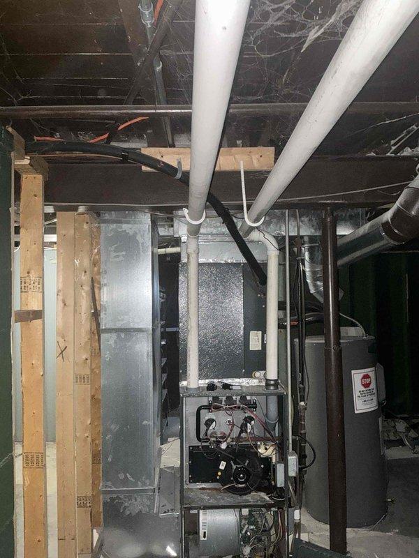 Conducted inspection of aging HVAC system in unfinished basement, documenting deteriorated ductwork, exposed wiring, and significant cobweb accumulation indicating poor airflow. Recommended complete system replacement due to observed inefficiencies, including compromised connections between concrete foundation and wooden joists where copper line set shows signs of wear.