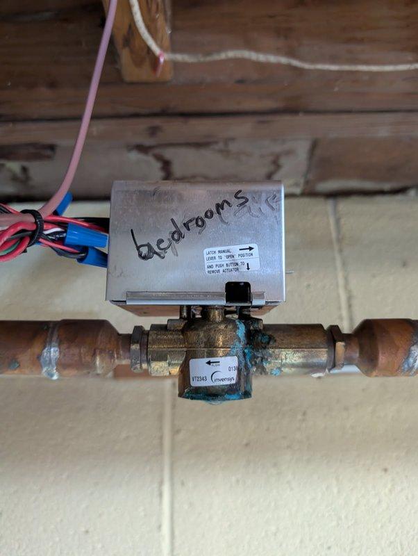 Performed zone valve inspection of hydronic heating system, identifying significant copper pipe corrosion at multiple connection points. Documented control valve with "bedrooms" zone designation and associated wiring (red, pink, blue, black) for control circuit. Noted extensive mineral buildup and oxidation at pipe joints, potentially indicating previous water leakage or ongoing moisture issues requiring future remediation.