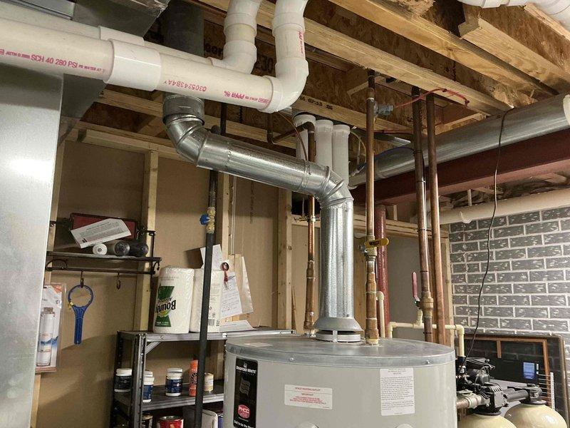 Conducted inspection of existing water heater system in basement utility area. Documented current installation which includes water heater tank with top connections, adjacent water treatment/filtration system, and associated plumbing infrastructure including copper pipes, PVC lines, and metal ducting. System evaluation completed for potential replacement of aging unit with more efficient model.
