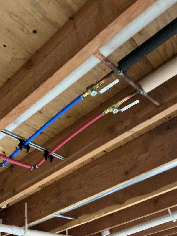 Completed installation of a PEX plumbing system in unfinished basement ceiling, including properly secured hot and cold water lines with brass valve fittings and white PVC drainage components. All pipes were properly mounted to wooden joists with appropriate brackets, ensuring a code-compliant installation with no leaks observed during final inspection.