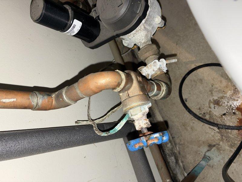 Conducted inspection of an aging water heater installation with significant copper pipe oxidation and mineral buildup at connection points. Documented existing plumbing configuration including copper/PVC pipe integration, valve assembly with prominent red handle, and pressure regulator system. Noted several areas requiring attention including corrosion at pipe junctions, deteriorating insulation, and potential flow restriction due to mineral deposits.