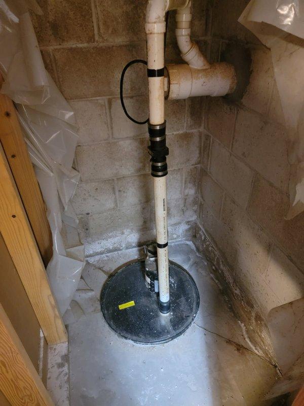 Conducted inspection of existing sump pump system in basement, documenting significant moisture intrusion and staining on cinder block foundation walls. Observed aging sump basin with visible sediment accumulation and corrosion on discharge piping. Recommended replacement of complete system including new basin, pump, check valve, and PVC discharge piping to prevent potential basement flooding.
