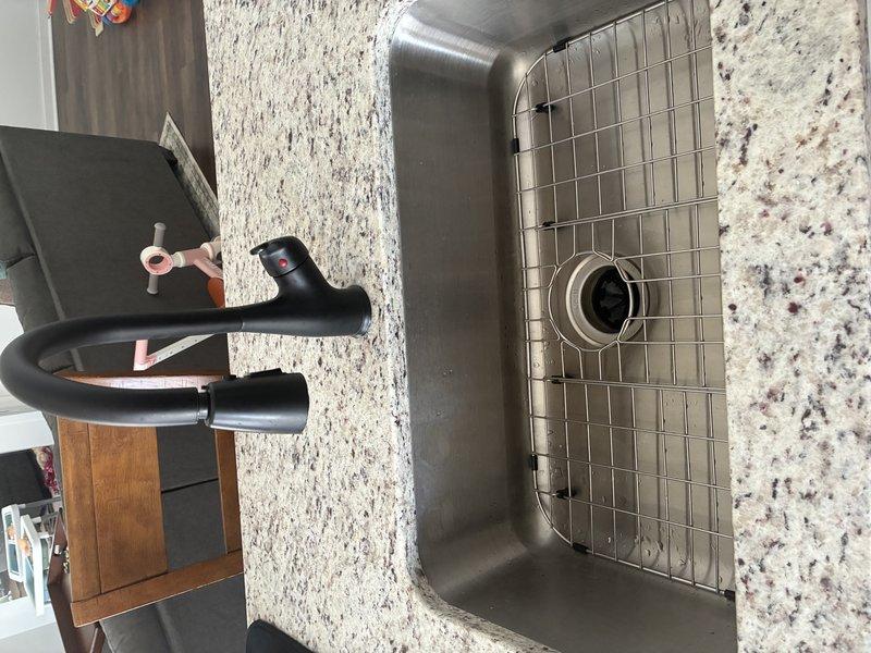 Installed new matte black kitchen faucet with pull-down sprayer head on granite/quartz countertop, connecting to existing supply lines. Secured all braided metal flexible hoses with proper metal clamps and verified connections to the blue cold water and red hot water supply pipes. System tested with no leaks observed.
