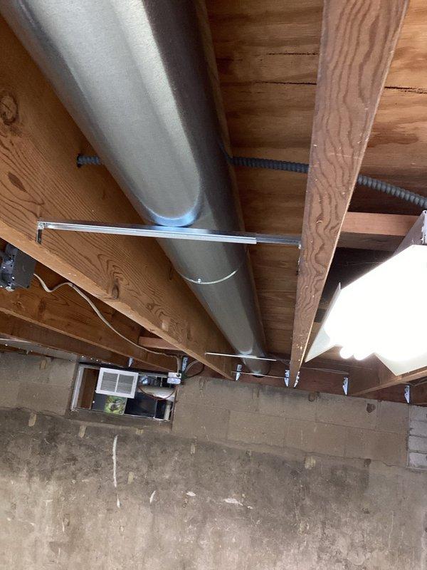 Installed cylindrical galvanized steel ductwork in basement/crawlspace area, securing the main trunk line to wooden joists with proper metal brackets and supports. The ductwork was successfully integrated with the existing infrastructure, running horizontally across the ceiling structure with connections to the ventilation system through the concrete block wall.