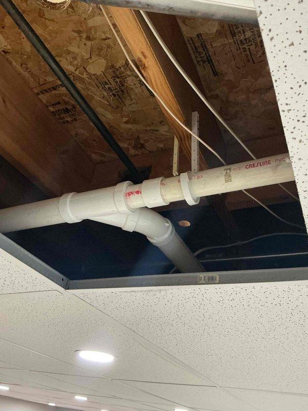 # Plumbing Install - Piping Job Description

Completed installation of white PVC piping system spanning ceiling cavity and interior wall framing. Routed Type CAE/LINE PVC pipe through overhead space above drop ceiling, utilizing proper fittings including T-joints and elbows to achieve required configuration. Secured piping to wooden joists with appropriate hangers and supports to prevent sagging and ensure code compliance.

Installed vertical PVC pipe runs through exposed wall framing, connecting to white plastic junction box housing red and blue valve controls. Configured complex routing pattern with multiple elbows and joints to navigate structural obstacles and achieve proper drainage slope. Integrated piping system through floor penetrations, ensuring proper sealing and support at transition points.

Mounted control valve assembly to wall studs at accessible height for future maintenance and operation. Verified all connections were properly solvent-welded and pressure-tested system