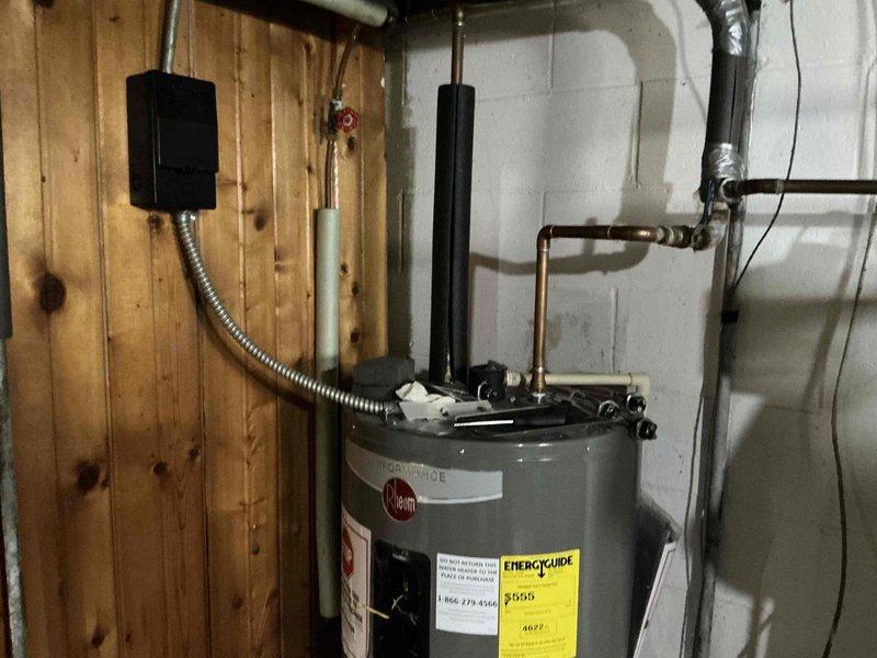 Completed HVAC estimate for system replacement in residential basement utility area. Assessed existing Rheem water heater installation with associated copper piping, electrical connections via junction box and flexible metallic conduit, and dual black vent pipes extending to ceiling. Evaluated secondary white HVAC unit with multiple copper pipe connections showing age-related oxidation, blue valve control point, flexible metallic ventilation duct, and associated wiring systems mounted on concrete block walls. Documented current equipment configuration, plumbing connections, electrical service, and venting arrangements to provide comprehensive replacement specifications and equipment sizing recommendations for complete system upgrade.