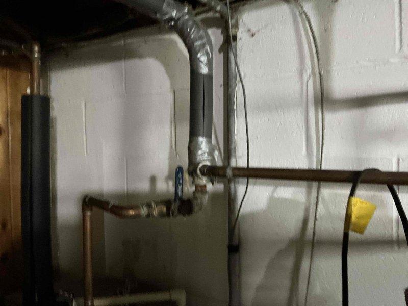 Completed HVAC estimate for system replacement in residential basement utility area. Assessed existing Rheem water heater installation with associated copper piping, electrical connections via junction box and flexible metallic conduit, and dual black vent pipes extending to ceiling. Evaluated secondary white HVAC unit with multiple copper pipe connections showing age-related oxidation, blue valve control point, flexible metallic ventilation duct, and associated wiring systems mounted on concrete block walls. Documented current equipment configuration, plumbing connections, electrical service, and venting arrangements to provide comprehensive replacement specifications and equipment sizing recommendations for complete system upgrade.