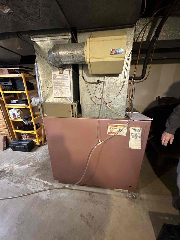 Conducted comprehensive evaluation of existing residential HVAC system located in basement utility area for equipment replacement estimate. Assessed current furnace unit, ductwork configuration, and associated mechanical components including water heater connections and ventilation systems. Documented equipment condition, spatial constraints, existing infrastructure including exposed ceiling joists, concrete flooring, and proximity to adjacent storage areas. Photographed current installation showing beige furnace with metallic ductwork, insulated ventilation components, electrical connections, and copper and PVC piping arrangements to provide detailed reference for replacement specifications and system upgrade recommendations.