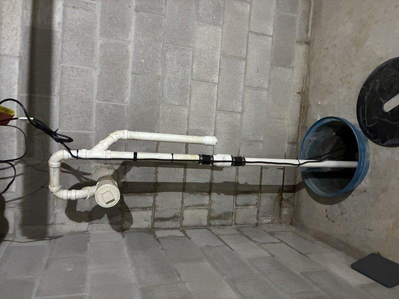 Completed sump pump installation in basement utility area, including new discharge piping system with white PVC drainage lines and proper floor drain connection. Installed copper piping assembly with ball valves (blue and red handles for easy identification) connected to the pump system, along with pressure relief components and necessary fittings to ensure proper operation and code compliance.