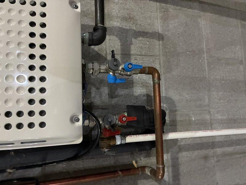 Completed sump pump installation in basement utility area, including new discharge piping system with white PVC drainage lines and proper floor drain connection. Installed copper piping assembly with ball valves (blue and red handles for easy identification) connected to the pump system, along with pressure relief components and necessary fittings to ensure proper operation and code compliance.