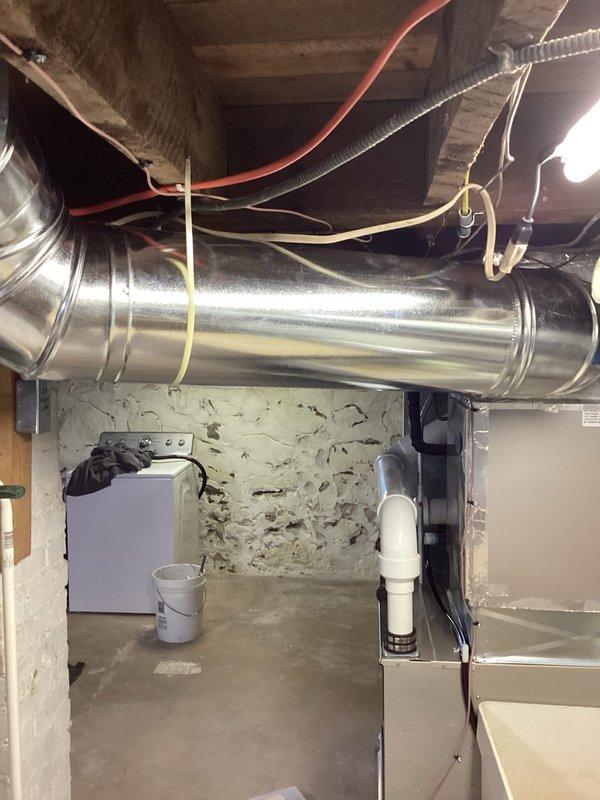 Completed HVAC system installation in residential basement utility area. Installed new beige cabinet furnace unit with dual white PVC intake and exhaust venting pipes routed vertically through ceiling structure. Configured sheet metal ductwork with angular transitions to integrate with existing air distribution system. Positioned white condensate pump adjacent to furnace on concrete floor for proper drainage management. Secured all metallic ductwork connections along ceiling joists with appropriate hangers and supports. Verified proper clearances from concrete block walls and ensured all venting, electrical connections, and drainage components meet manufacturer specifications and local code requirements. System tested and operational upon completion of installation warranty work.