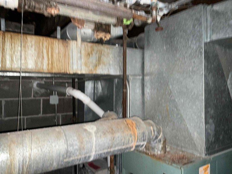 Completed comprehensive assessment of aging HVAC system in commercial mechanical space for equipment replacement estimate. Evaluated existing turquoise cabinet-style furnace showing significant deterioration, faded exterior finish, exposed internal components, and accumulated dust buildup indicating end of serviceable life. Documented large metal ductwork, insulated flexible ducts, and associated piping infrastructure throughout mechanical room, noting surface corrosion on overhead pipes and overall system condition. Provided detailed photographic documentation and replacement recommendations for outdated heating and air distribution equipment to improve efficiency and reliability.
