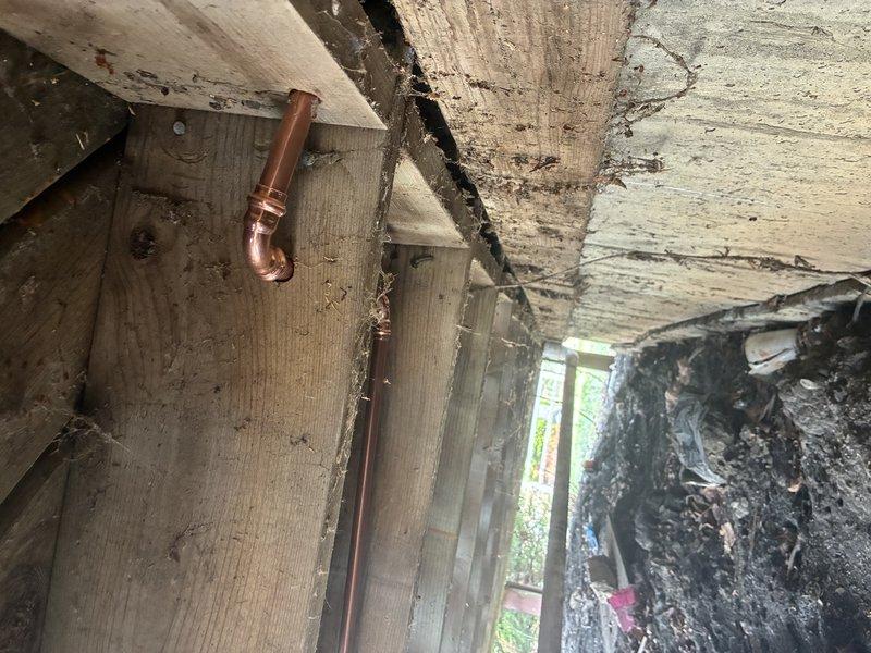 Responded to service callback to inspect and address plumbing concerns in deteriorated upper-level structure. Upon arrival, discovered exposed copper supply line running vertically through compromised ceiling area with significant structural deterioration, including extensive paint failure, water damage, and charred surfaces indicating previous fire damage. Accessed attic space through ceiling opening to evaluate copper piping installation and verify proper support and insulation clearances around pipe penetrations through floor joists. Confirmed copper line routing and connections were secure despite surrounding structural compromise. Documented existing conditions showing fiberglass insulation placement around framing members and pipe penetrations for reference.