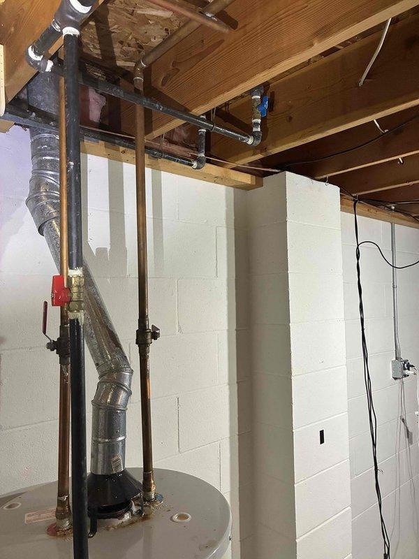 Completed water heater replacement in basement utility area. Removed existing unit and installed new cylindrical water heater tank with proper venting system, including galvanized metal ductwork with elbow joint extending through ceiling joists. Connected copper supply lines, gas piping with shut-off valve, and integrated all plumbing connections to existing infrastructure, securing pipes to wooden joists with appropriate strapping and supports.