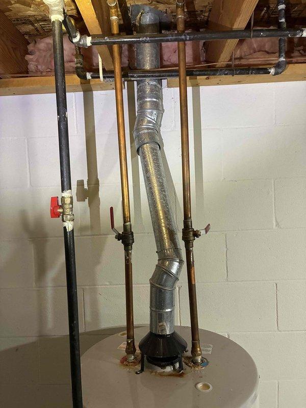 Completed water heater replacement in basement utility area. Removed existing unit and installed new cylindrical water heater tank with proper venting system, including galvanized metal ductwork with elbow joint extending through ceiling joists. Connected copper supply lines, gas piping with shut-off valve, and integrated all plumbing connections to existing infrastructure, securing pipes to wooden joists with appropriate strapping and supports.