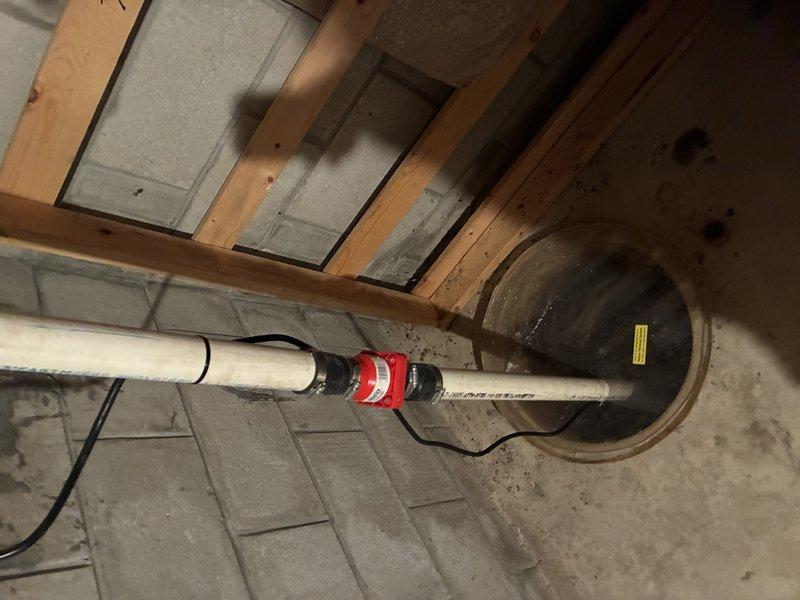 Performed sump pump inspection and documentation in residential basement utility space. Examined existing sump pump system including discharge piping with PVC connections, pump basin condition, and operational components. Basin showed significant sediment buildup, mineral deposits, and water level staining on interior walls, indicating regular water accumulation and pump cycling. Documented pump housing condition, discharge pipe configuration with visible red coupling connection, and drainage system including corrugated intake line. System components appeared functional with proper floor drain cover installation and secure pipe mounting hardware in place.