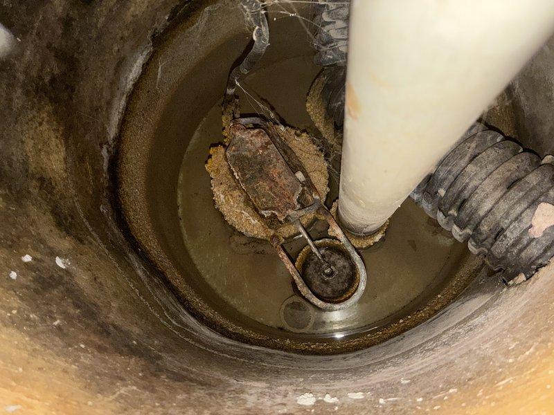 Performed sump pump inspection and documentation in residential basement utility space. Examined existing sump pump system including discharge piping with PVC connections, pump basin condition, and operational components. Basin showed significant sediment buildup, mineral deposits, and water level staining on interior walls, indicating regular water accumulation and pump cycling. Documented pump housing condition, discharge pipe configuration with visible red coupling connection, and drainage system including corrugated intake line. System components appeared functional with proper floor drain cover installation and secure pipe mounting hardware in place.