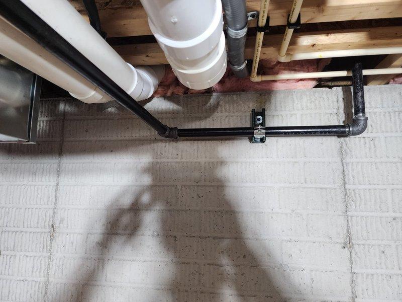 Installed hanging heater unit in basement area with exposed floor joists and existing utility infrastructure. Positioned and mounted heater to accommodate multiple existing plumbing lines including PVC drainage pipes, black waste lines, and gray flexible ductwork while maintaining proper clearances from wooden structural members and insulation. Secured unit with appropriate mounting hardware to ensure stable installation in unfinished below-grade space with concrete foundation.