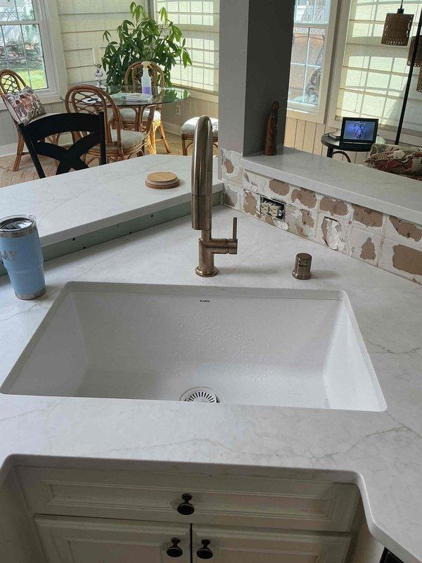 Completed plumbing estimate for kitchen fixture installation featuring an undermount sink with gold-finished faucet, matching soap dispenser, and garbage disposal system. Assessed existing white cabinetry configuration with proper drainage connections including white PVC piping and functional disposal unit with electrical hookup beneath sink area.