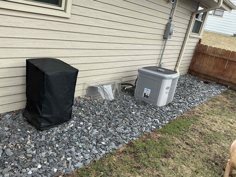 Completed HVAC system replacement at residential property, installing new Carrier Puron heat pump condenser unit on prepared gravel pad against exterior wall. Unit was positioned with proper clearances, electrical conduit and disconnect box were mounted to building siding, and all connections were secured according to manufacturer specifications and local code requirements.