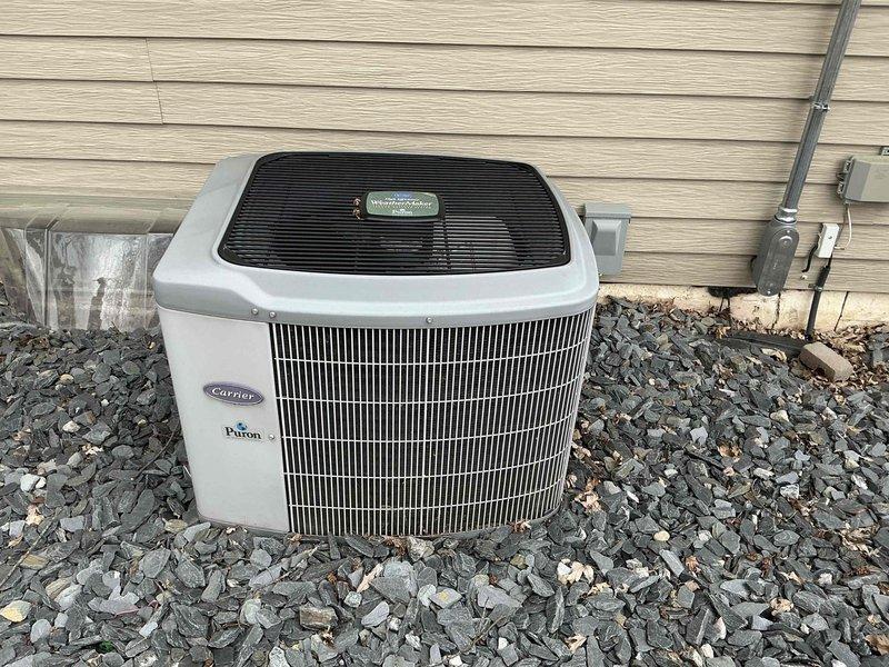Completed HVAC system replacement at residential property, installing new Carrier Puron heat pump condenser unit on prepared gravel pad against exterior wall. Unit was positioned with proper clearances, electrical conduit and disconnect box were mounted to building siding, and all connections were secured according to manufacturer specifications and local code requirements.