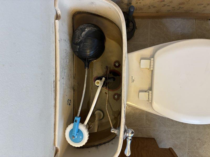 Responded to commercial toilet maintenance call and performed internal tank inspection and component assessment. Removed tank lid to evaluate flushing mechanism, which revealed significant mineral buildup and discoloration throughout tank interior indicating extended service life. Inspected float ball assembly with plastic arm mechanism and blue base component for proper operation. Assessed tank condition showing brown water staining and mineral deposits concentrated around waterline and mechanical connection points. Documented existing flush valve system and chrome flush handle operation. Tank components showed age-related wear but remained functional. Verified toilet positioning and surrounding fixtures in commercial restroom environment with proper mounting and stability.